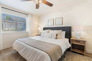 Une chambre à coucher avec un grand lit, deux tables de chevet, un ventilateur de plafond et une fenêtre avec des stores.