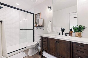 Une salle de bain moderne avec une douche à parois de verre, des toilettes blanches, un meuble-lavabo en bois foncé agrémenté d'un miroir et une petite étagère avec des objets décoratifs.