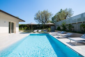 Una piscina rectangular con agua azul cristalina, rodeada de sillas reclinables y sombrillas blancas, con árboles y un edificio moderno al fondo.
