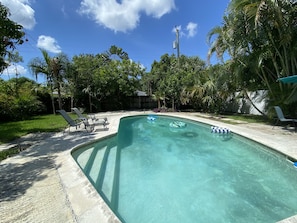 Une piscine aux eaux cristallines, bordée de chaises longues et de bouées gonflables, entourée d’une végétation luxuriante et sous un ciel dégagé.