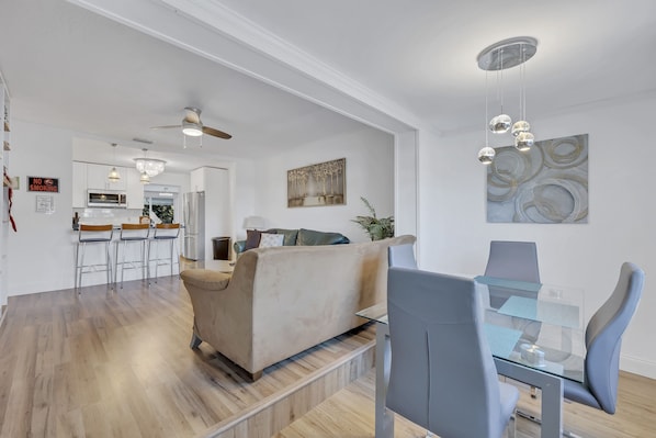 Un salon moderne avec une table à manger en verre, des chaises grises, un canapé beige et une cuisine en arrière-plan.