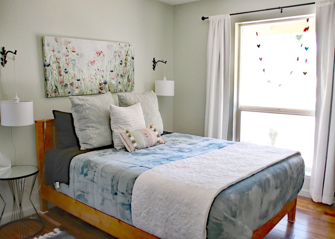 Quarto com cama, abajures, janela com cortinas e um quadro com flores na parede.