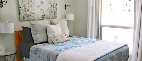 Quarto com cama, abajures, janela com cortinas e um quadro com flores na parede.