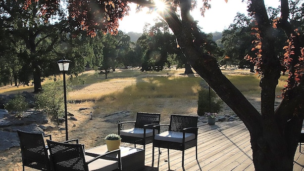 A wooden deck with two wicker chairs and a table, surrounded by trees and a lamp post. - Private Home at the foot of Yosemite National Park (Mariposa)