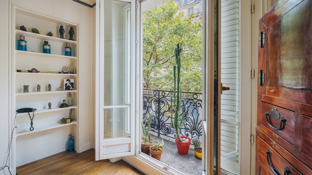 A room with a wooden floor, white shelves with various items, a wooden cabinet, and a balcony with potted plants and a metal railing. - Large 1BR close to Bastille and Aligre (Paris)
