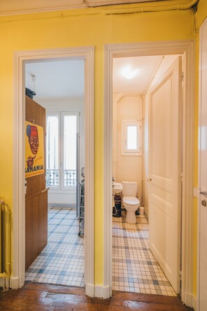 A narrow hallway with a tiled floor, leading to a bathroom with a toilet and a window. - Large 1BR close to Bastille and Aligre (Paris)