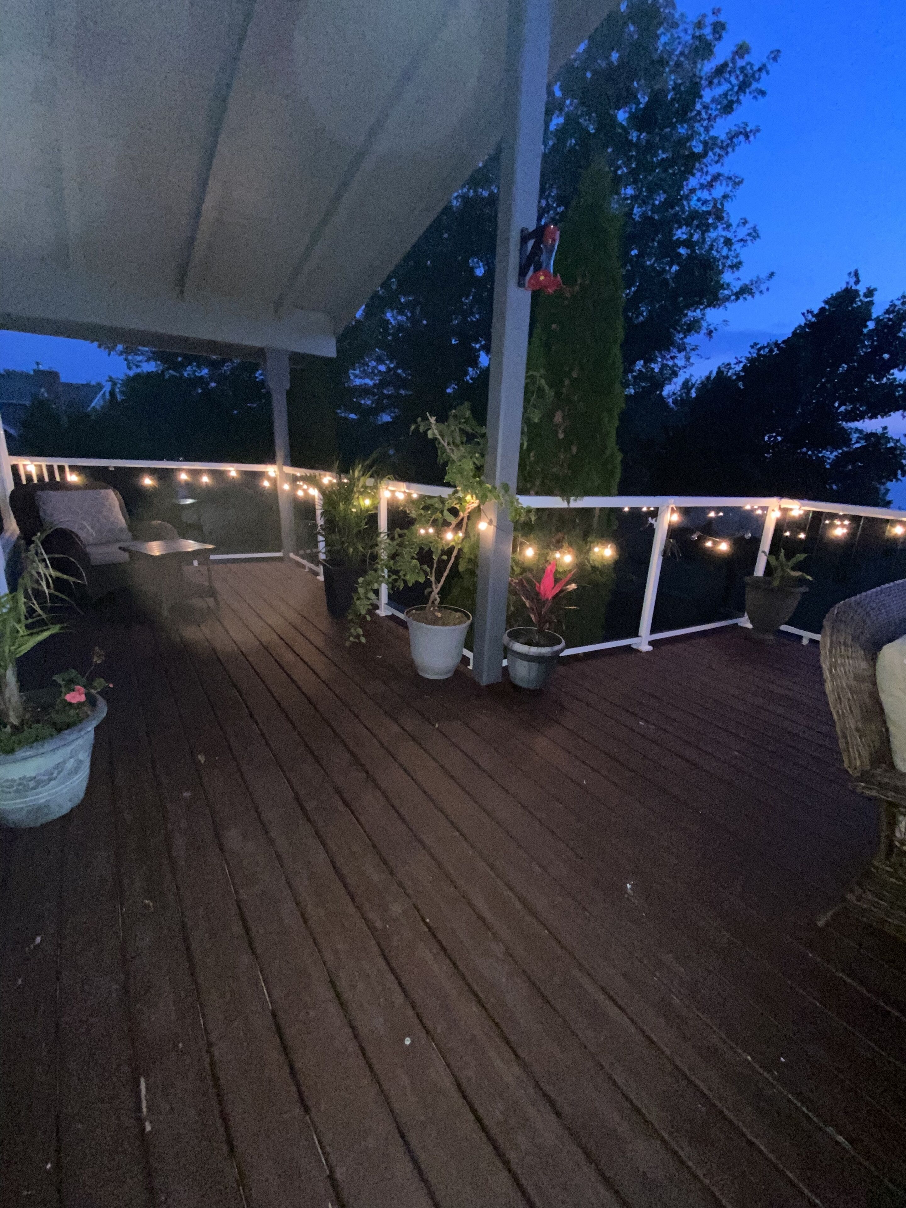 Una terraza de madera con luces colgantes, plantas en macetas y mobiliario exterior.