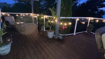 Una terraza de madera con luces colgantes, plantas en macetas y mobiliario exterior.