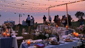 Un área de comedor al aire libre con luces colgantes y mesas preparadas para una comida, con vista a un cuerpo de agua.