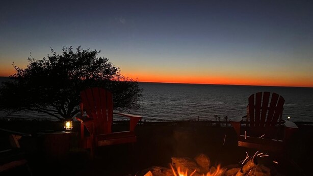Una chimenea con dos sillas Adirondack y un farol, con vista a la puesta de sol sobre el océano.