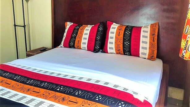 A bed with a patterned bedspread and pillows, a wooden headboard, and a floor lamp. - Modern studio decorated in African colors at the rear of a spacious villa. (Yaoundé)