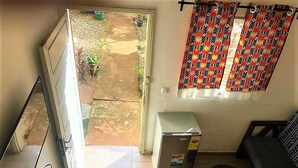 A compact living space with a sliding door leading to a garden, a small refrigerator, a sofa, and a black trash bin. - Modern studio decorated in African colors at the rear of a spacious villa. (Yaoundé)