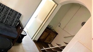 A narrow hallway with a tiled floor, a black toilet, a patterned bench, and a doorway leading to a small room with a wooden cabinet. - Modern studio decorated in African colors at the rear of a spacious villa. (Yaoundé)