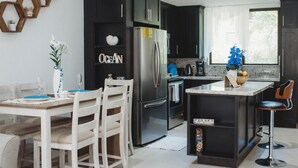Una cocina moderna con armarios oscuros, un refrigerador de acero inoxidable, una zona de comedor con sillas blancas y un taburete de bar.