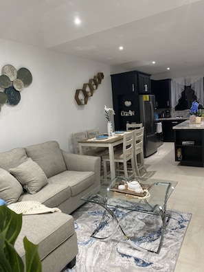 A modern living room with a grey sofa, a glass coffee table, and a dining area with a wooden table and chairs. - JeSal’s Escape 3 Bedroom Townhome Negril- 5 min From Beach- Perfect For Longterm (Negril)