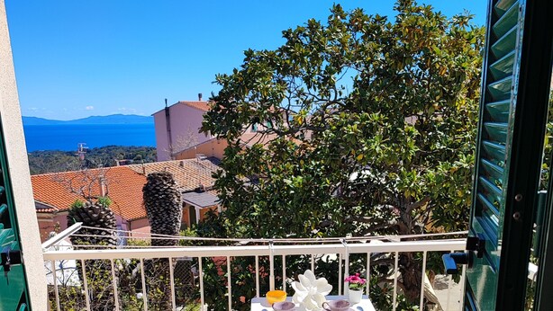 A balcony with two white metal chairs, a small table with a flower vase, and a view of the sea and mountains.