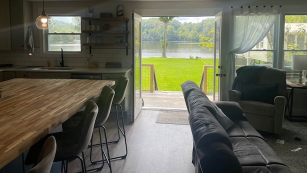 A modern kitchen with a large wooden table and chairs, a built-in shelf with glassware, and a view of a lake through open doors.