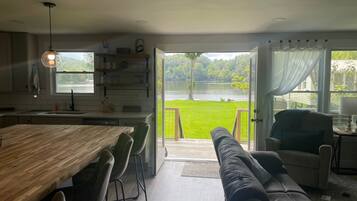 Een moderne keuken met een grote houten tafel en stoelen, een inbouwkast met glazen en een uitzicht op een meer door open deuren.