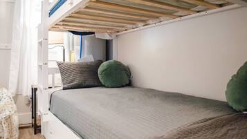 Uma cama beliche com colchão cinza e duas almofadas, uma verde e outra listrada, num quarto com paredes brancas e uma janela.