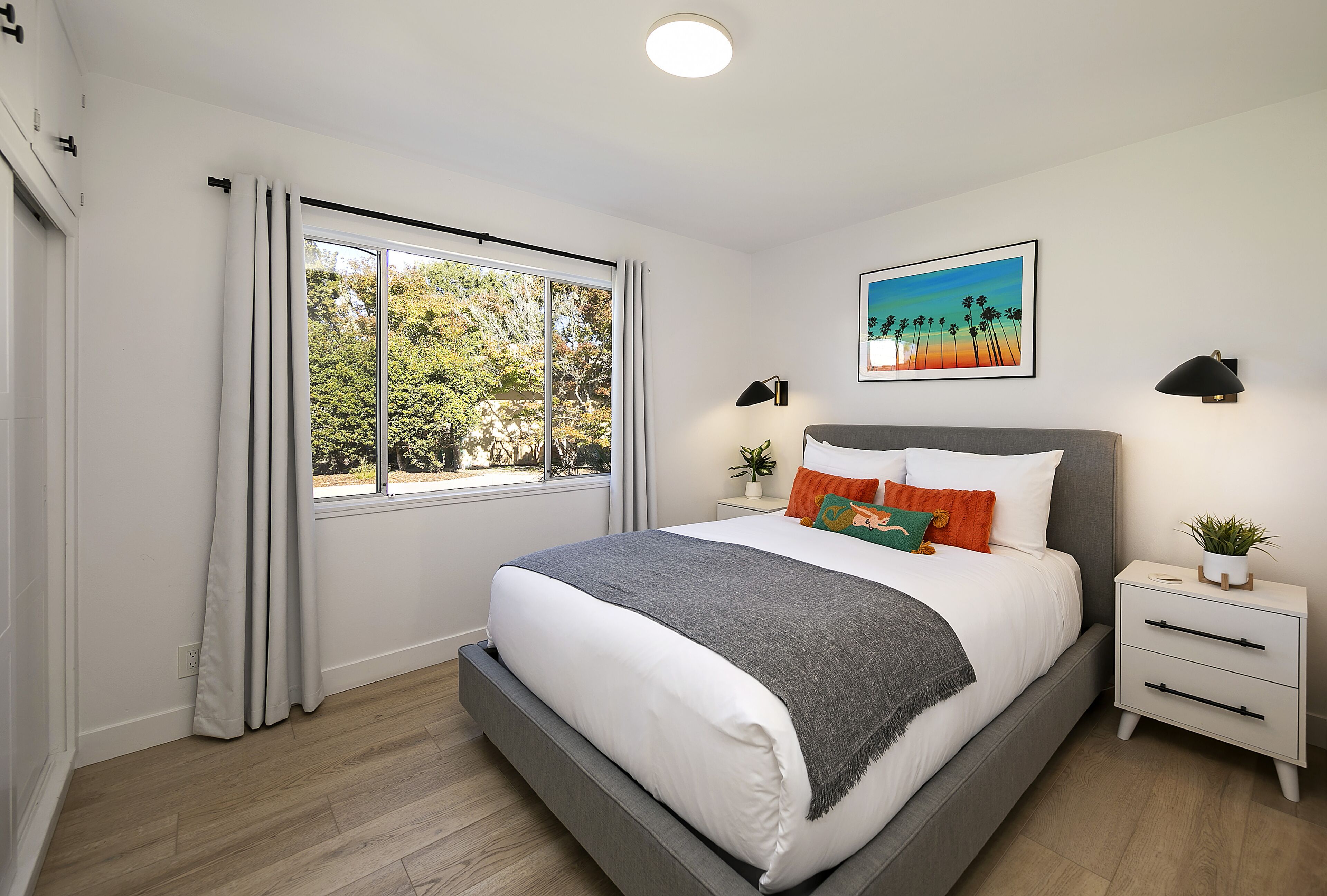A modern bedroom with a large bed, a nightstand, a window with curtains, a wall-mounted lamp, and a framed picture.