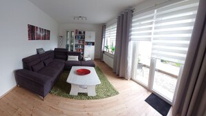 Ein Wohnzimmer mit einem Ecksofa, einem weißen Couchtisch mit einer roten Schüssel, einem Bücherregal und Blick ins Freie durch große Fenster mit Jalousien.