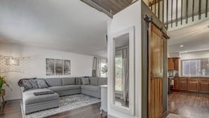 A modern living room with a grey sectional sofa, a small ottoman, and a patterned rug. A large mirror reflects the room, and a sliding barn-style door leads to the kitchen. - Inviting 5 Bedroom Home Centrally Located (Colorado Springs)