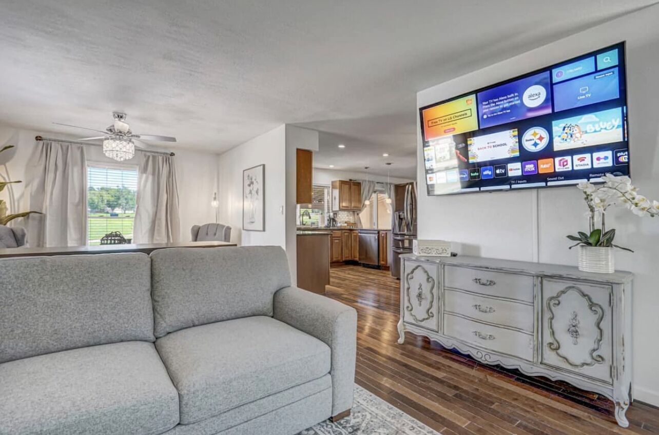 Un salon avec un canapé gris, un meuble blanc et un téléviseur à écran plat fixé au mur, qui diffuse différentes chaînes.
