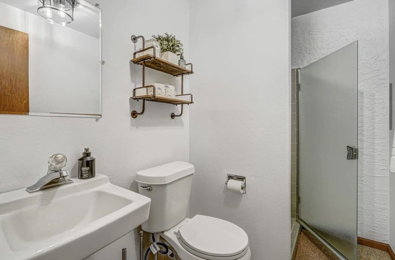 Et badeværelse med en hvid vask, toilet og en brusekabine med glasvægge. Træhylder med toiletartikler og en lille plante.