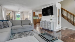 A modern living room with a grey sofa, a flat-screen TV mounted on the wall, a wooden staircase, and a kitchen in the background. - Inviting 5 Bedroom Home Centrally Located (Colorado Springs)