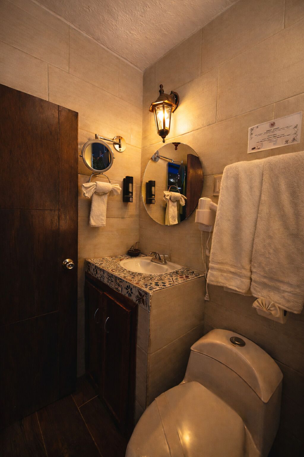 A bathroom with a wooden door, a round mirror, a sink with a patterned tile backsplash, a toilet, and a towel rack.
