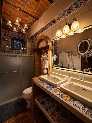Une salle de bain avec un mur recouvert de carreaux, un grand miroir, deux lavabos, des toilettes et un espace douche.