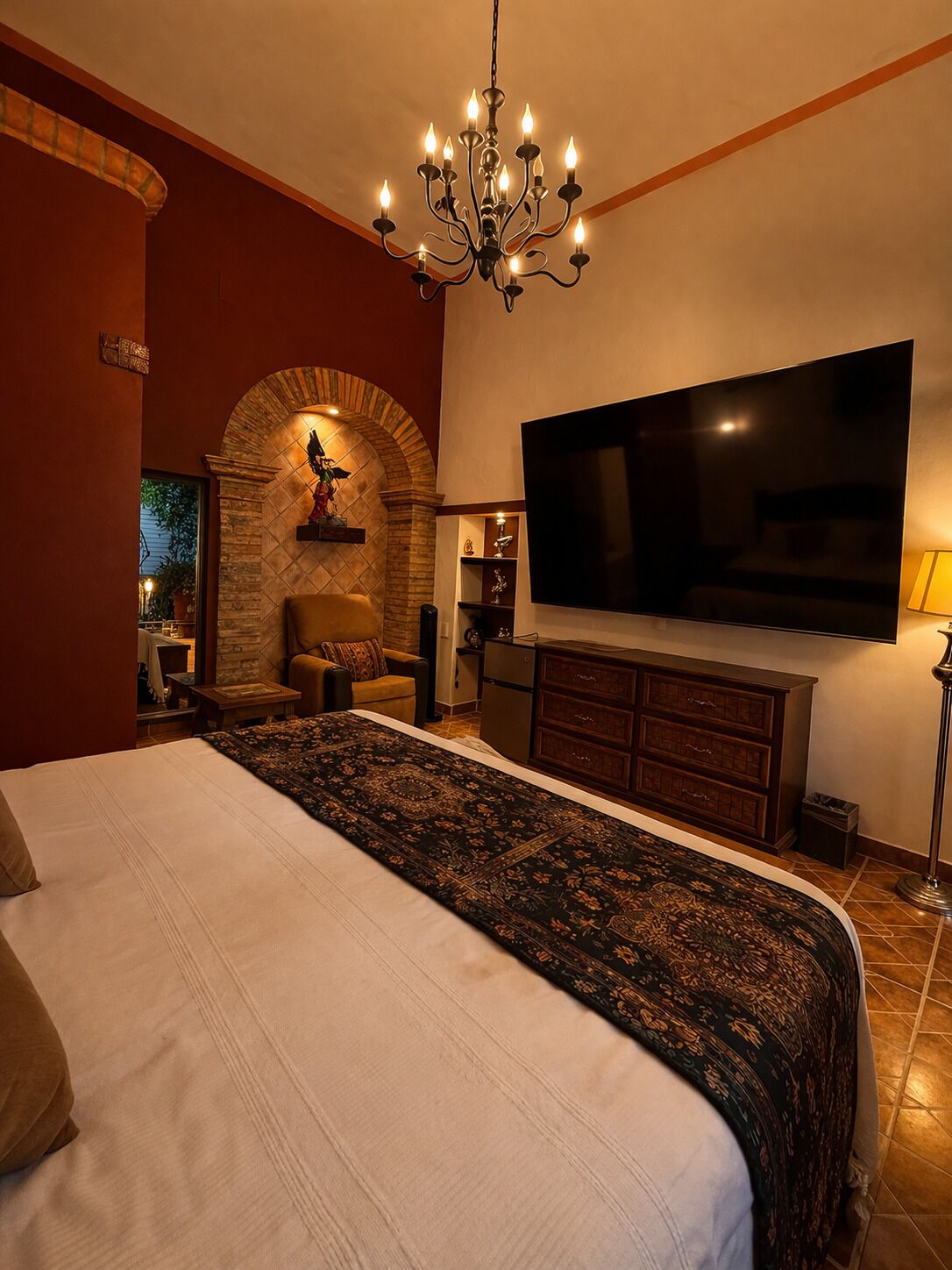 A bedroom with a large bed, a TV mounted on the wall, a chandelier, and a fireplace.