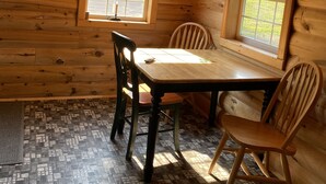 A wooden cabin interior with a dining table, four chairs, two windows, and a tiled floor.