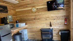 A compact cabin-style room with a wooden interior, a clock, a television, a microwave, a refrigerator, a trash bin, and a small heater.