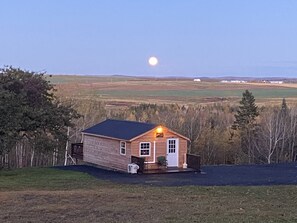 Una cabaña de madera con un porche, un cobertizo pequeño y una vista despejada de la luna en el cielo.