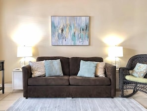 A living room with a brown sofa, two side tables with lamps, a wicker chair, and a painting on the wall.