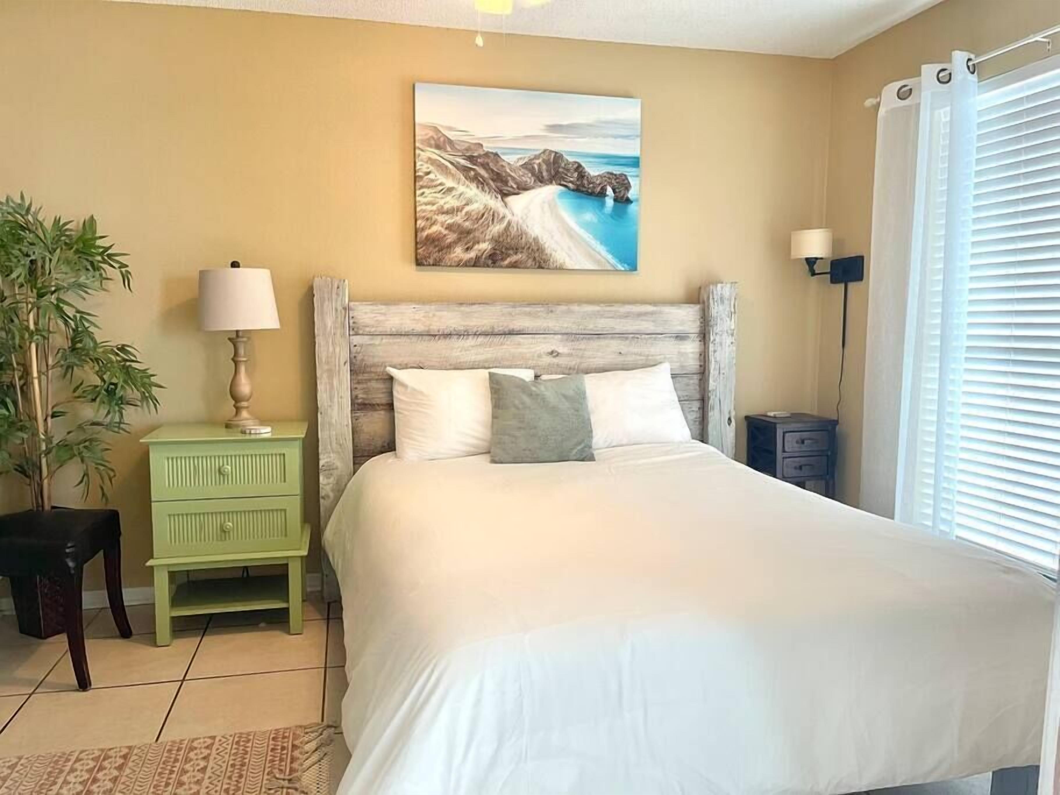 Quarto com cama, mesa de cabeceira, abajur, planta em vaso e um quadro na parede.