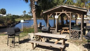 Un belvédère en bois, équipé de tables de pique-nique et d’un barbecue, offre une vue sur un plan d’eau où l’on aperçoit des bateaux et des maisons au loin.
