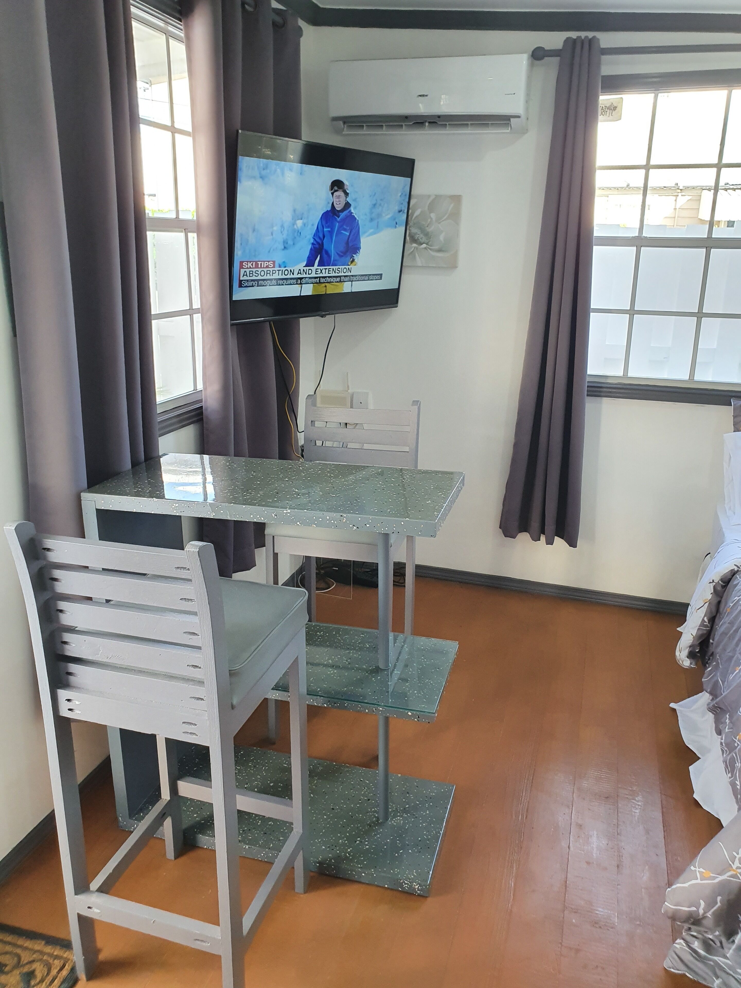 A room with a TV mounted on the wall, a table with chairs, and a window with curtains.
