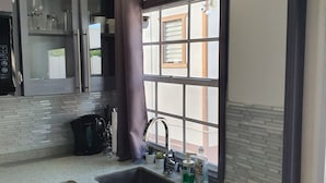 A kitchen with a stainless steel sink, a window with blinds, and a black curtain. - Devachan Apartments (Worthing)
