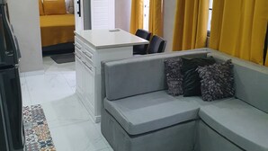A compact living space with a grey sofa, a small kitchenette, and a bed with yellow bedding. - Devachan Apartments (Worthing)