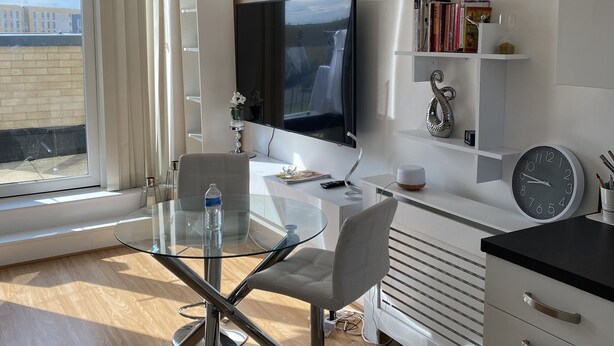 Una sala de estar moderna con una mesa de comedor de cristal, un televisor montado en la pared y una estantería con libros y objetos decorativos.