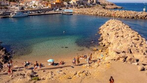 Una playa costera con terreno rocoso, agua cristalina y edificios al fondo.