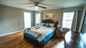 Une chambre avec un plancher en bois, un lit recouvert d’une courtepointe blanche et bleue, une table de chevet, une commode et un ventilateur de plafond.