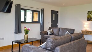 A living room with a sofa set, a small table, a TV mounted on the wall, a window with curtains, and a door. - Offa's Dyke Lodge - UK10835 (Stanner, near Kington)