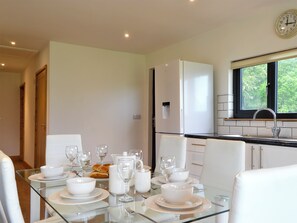 A dining area with a glass table set for a meal, white chairs, a white refrigerator, and a window with a view of greenery.