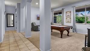 A spacious hallway with a pool table, a comfortable chair, and a large window offering a view of the outdoors.