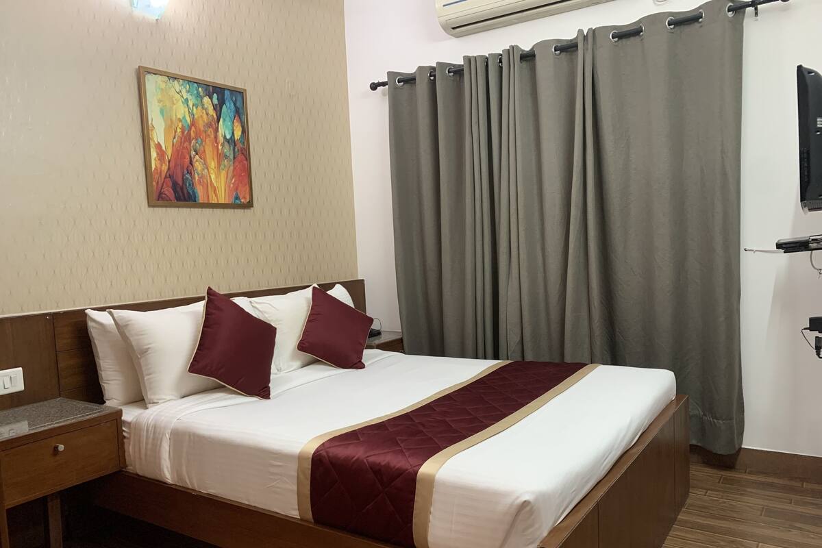 Cama bem arrumada com roupa de cama branca e bordô, cabeceira de madeira, mesa de cabeceira, um quadro na parede e cortina cinza.