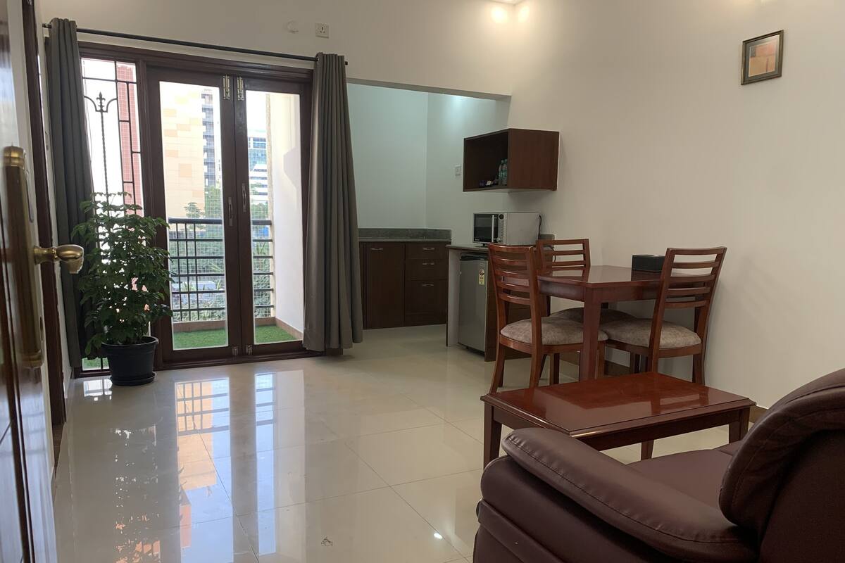Uma sala de estar moderna com sofá, mesa de jantar com cadeiras, área de cozinha com micro-ondas e uma porta de vidro deslizante que dá para uma varanda.