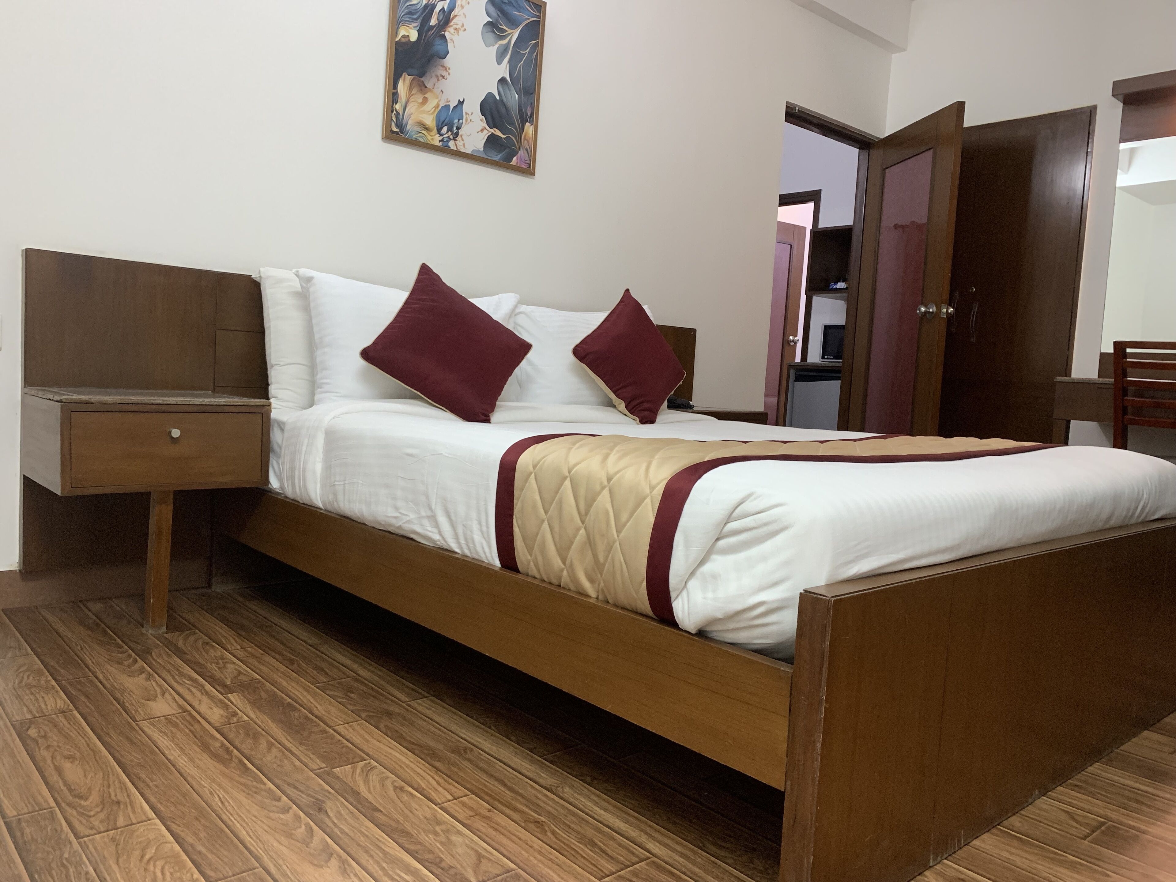 A neatly made bed with a wooden frame, white bedding, and two red pillows. There is a wooden nightstand with a drawer to the left. A wooden door is visible in the background.